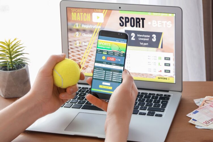 Picture of a man holding surfing a sports betting website in his mobile and laptop while handling a tennis ball.